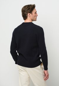 Marinblå stickad tröja med ribbad textur, rund halsringning och långa ärmar, i kombination med off-white byxor, sedd bakifrån.