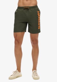 Mann trägt olivgrüne Badeshorts mit orangefarbenem "Superdry"-Logo auf dem rechten Bein und weiße Leinenschuhe, steht vor weißem Hintergrund.
