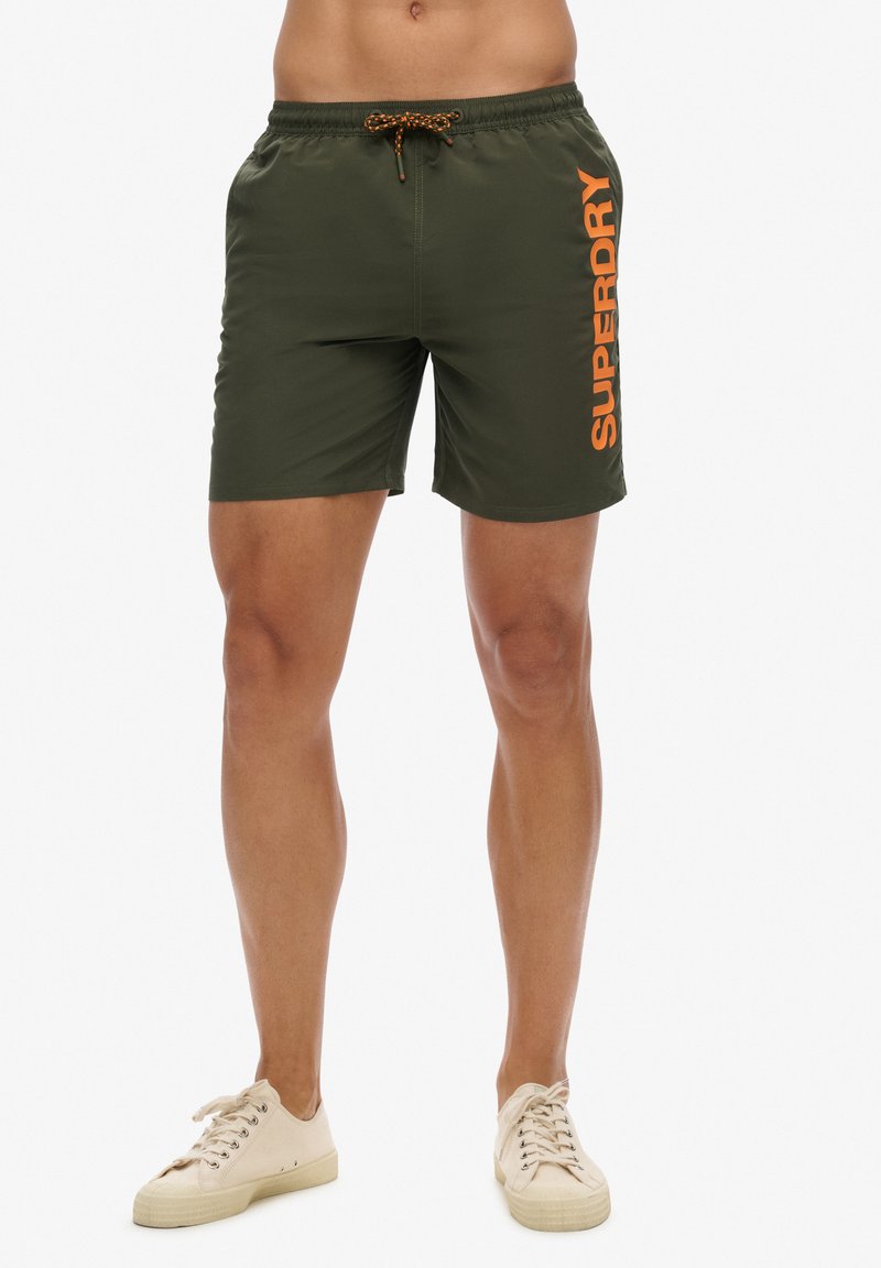 Mann trägt olivgrüne Badeshorts mit orangefarbenem "Superdry"-Logo auf dem rechten Bein und weiße Leinenschuhe, steht vor weißem Hintergrund.