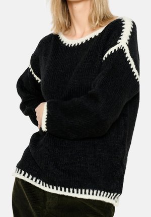 Femme portant un pull noir en maille avec des bords blancs cousus au niveau de l'encolure, des épaules, des poignets et de l'ourlet, assorti à un pantalon vert foncé.