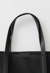 Zwarte tote bag van gaas materiaal met gladde leren handvatten. Heeft een rechthoekige vorm en een merklabel aan de voorkant.