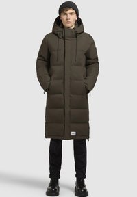 Lang, polstret parka i mørk olivengrøn med hætte, lynlåslommer og et logo-patch, båret over mørke bukser og sorte støvler.