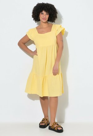 Kobieta z kręconymi włosami stoi, ubrana w żółtą sukienkę do kolan i sandały w panterkę na jednolitym białym tle.