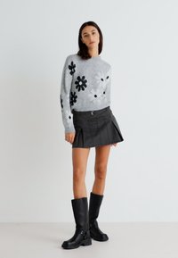 Grauer Pullover mit schwarzen Blumenmustern, kombiniert mit einem schwarzen Faltenrock und kniehohen schwarzen Stiefeln mit Schnallen.