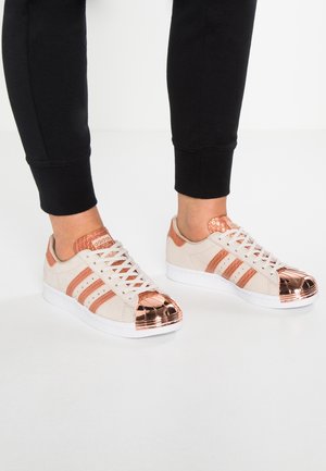 Pieds portant des baskets Adidas beige avec embouts et rayures en or rose métallique, associées à un pantalon noir, debout sur un sol blanc.