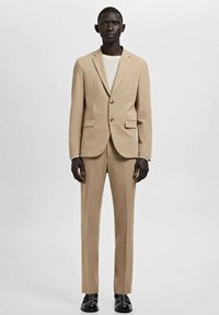 Traje beige a medida hecho de tela texturizada, con blazer de un solo botón, dos bolsillos delanteros y pantalones a juego. Zapatos negros complementan el conjunto.
