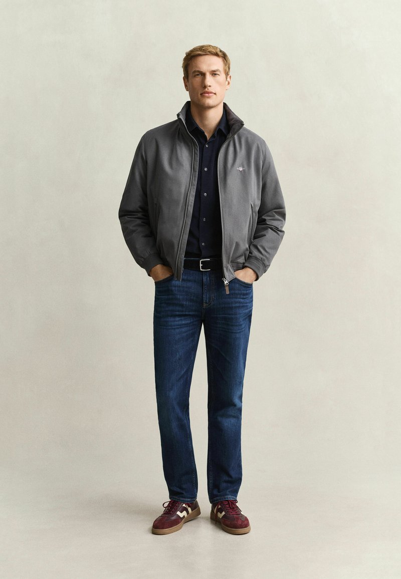 Man standing with hands in pockets wearing a grey zip-up jacket, dark shirt, blue jeans, and maroon sneakers against a plain background.