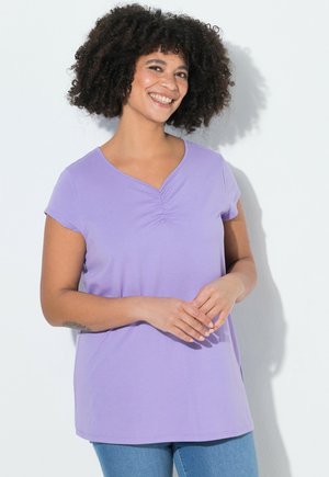 Femme aux cheveux bouclés portant un haut à col en V lavande froncé et un jean bleu, souriante, les mains doucement jointes devant elle.