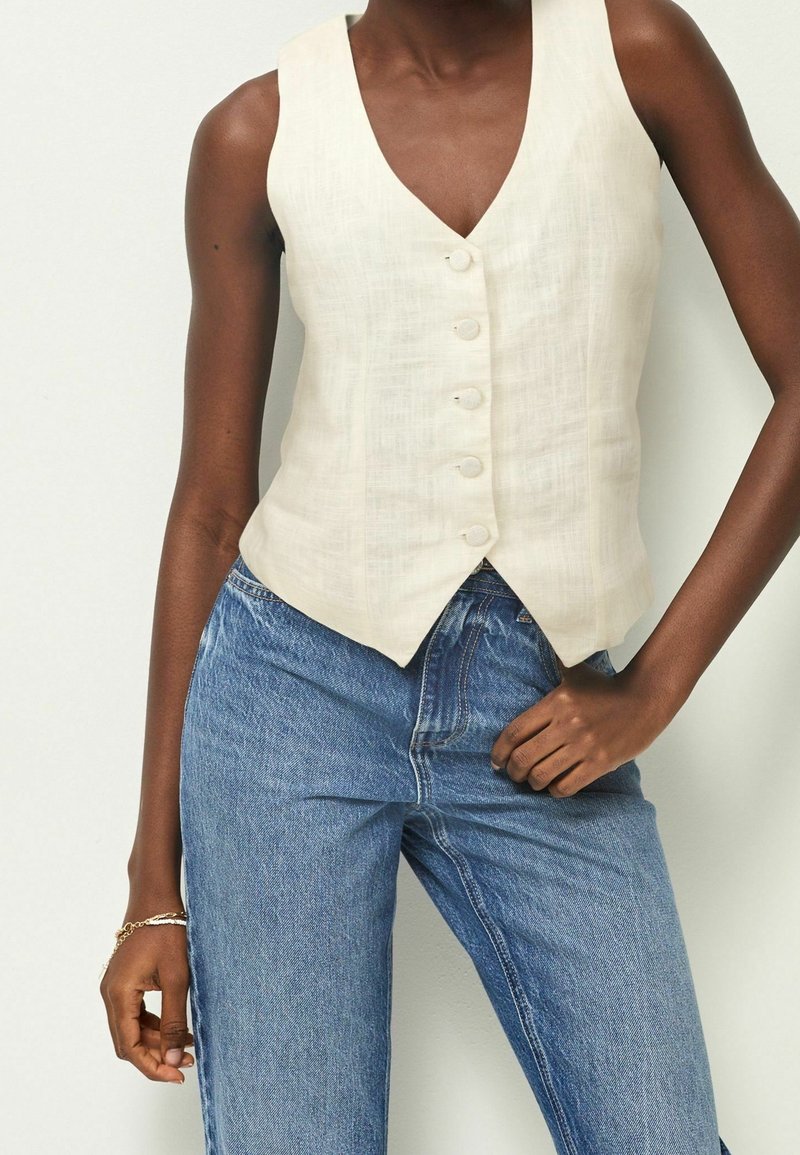Linnen mouwloos vest, crème kleur, V-halsontwerp met vijf knoopsluitingen. Gecombineerd met lichtblauwe denim jeans. Glad textuur en slim fit stijl.