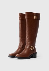 Une paire de bottes en cuir brun, hautes, avec des boucles en or sur des talons bas noirs, posée sur un fond blanc uni.