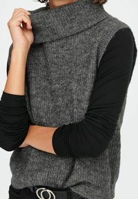 Personne portant un pull en maille grise sans manches avec un col roulé par-dessus un haut à manches longues noir, avec une main près du visage et les bras croisés.