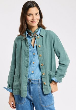 Femme portant une veste verte boutonnée sur une chemise en jean et un jean, avec une écharpe verte nouée autour du cou, souriant les mains dans les poches.