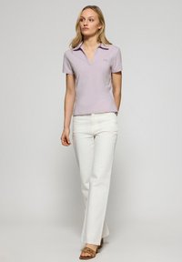 Jasnofioletowa koszulka polo z dekoltem w serek, krótkimi rękawami i subtelnym detalem z logo, zestawiona z białymi spodniami o szerokich nogawkach i brązowymi sandałami.