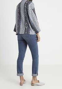 Blouse à manches trois-quarts, ample et à motifs en bleu et gris, portée avec un jean ajusté à poignets retroussés et des chaussures légères.