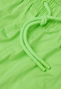 Neon zelené plavky s texturovaným povrchem, s upleteným šňůrkovým páskem a plastovými konci, plus šité detaily na látce.