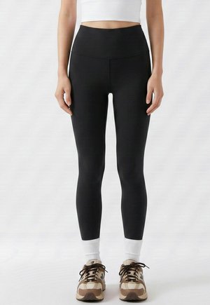 Femme portant des leggings noirs taille haute, un top court blanc, des chaussettes blanches et des baskets beige et marron, debout devant un fond uni.