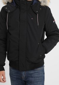 Černá bunda s kapucí lemovanou kožešinou, na zip vpředu, s elastickými manžetami a šňůrkami jako zdobení. Texturovaná tkanina s bočními kapsami. Neformální střih.