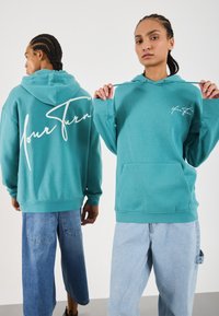 Dos personas con cabello trenzado que llevan sudaderas azuladas; una se enfrenta sosteniendo los cordones, y la otra muestra un gran texto blanco en la parte de atrás.