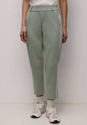 Osoba ubrana w jasnooliwkowe spodnie z fakturą, z białymi paskami po bokach, białe skarpetki i białe masywne sneakersy, stojąca na jednolitym tle.