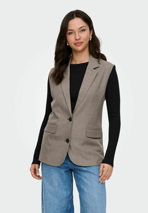 Femme portant un blazer sans manches beige sur un top noir à manches longues et un jean bleu, debout devant un fond clair et uni.