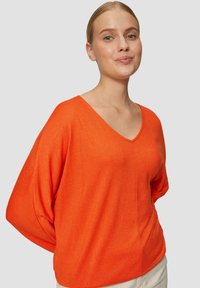 Helle orange V-Ausschnitt-Sweater mit lockerem Schnitt und Fledermausärmeln. Glatte Textur und minimalistische Details, kombiniert mit hellen Hosen.