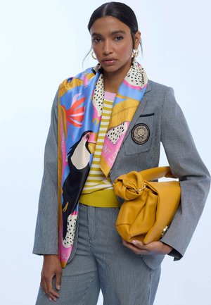 Bufanda de seda con patrones coloridos de animales y plantas, drapeada sobre un blazer gris a rayas. La modelo sostiene un bolso de cuero amarillo.