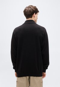 Μαύρο πλεκτό πουλόβερ με ριμπ γιακά και μανσέτες, με άνετη εφαρμογή. Το ύφασμα φαίνεται απαλό και έχει λεία υφή.