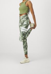 Oliwkowy crop top o dopasowanym kroju w połączeniu z leginsami o wysokim stanie, które mają zielony tropikalny wzór oraz białe pasy akcentujące.