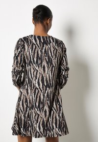 Donna con i capelli raccolti a chignon, vestita con un abito a maniche lunghe e lunghezza al ginocchio, caratterizzato da strisce astratte a forma di zebra nei colori nero, marrone e beige, davanti a un muro bianco.