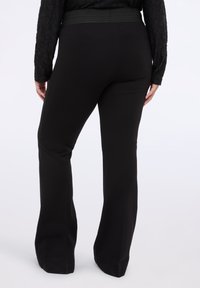 Černé kalhoty s rozšířenými nohavicemi a žebrovaným pasem, vyrobené z hladkého materiálu. Pohled zezadu ukazuje přiléhavou siluetu, která se rozšiřuje do zvonového lemu.