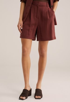 Nő magas derekú, bő fazonú rozsda színű rövidnadrágban és fekete szövött ék sarkú szandálban, kezét részben a zsebében tartva.