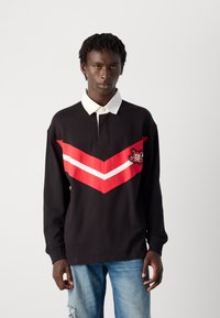 Giovane con capelli neri intrecciati, che indossa una maglietta da rugby nera con colletto bianco, strisce a zig zag rosse e bianche, e jeans strappati azzurri.