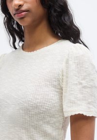 Φωτεινό μπεζ, υφασμένο τοπ με κοντά φουσκωτά μανίκια και τελείωμα με κυματιστό κόψιμο. Κατασκευασμένο από μαλακό, διαπνέον ύφασμα με διακριτικές κάθετες γραμμές.