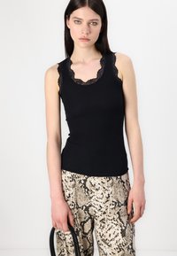 Débardeur noir à rayures avec un col en dentelle festonné, associé à un pantalon à imprimé serpent beige et noir. Le mannequin tient un sac à main noir.