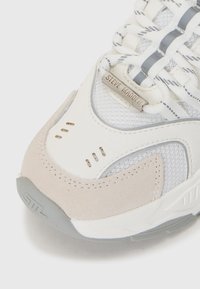 Baskets présentant un mélange de cuir blanc et de daim gris, des panneaux en mesh, des lacets fins et une étiquette en métal gravée "Steve Madden."