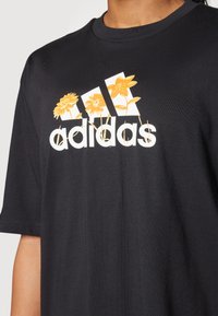 Black cotton t-shirt featuring a white adidas logo with yellow floral accents above it. Short sleeves and a crew neckline.