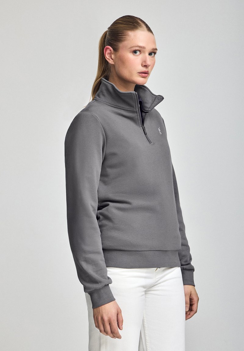 Grå zip-up sweatshirt med hög krage, ribbade ärmslut och nederkant, tillverkad av mjukt material, med en liten logotyp på bröstet, bärs med vita byxor.