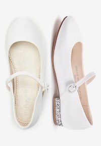 Białe buty typu Mary Jane, jeden pokazujący widok z góry z paskiem i falistą wkładką, a drugi widok z boku z błyszczącym niskim obcasem.