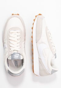 Nike-sneakers i beige och ljusgrått, med ovandel i tyg, mockadetaljer, platta skosnören och en gummisula med framträdande mönster.