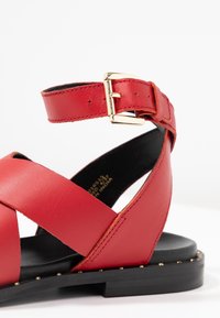 Punaiset nahkasaappaat, joissa on ristiin menevät hihnat, kultainen solki ja musta pohja, jossa on pieniä kultaisia niittejä reunoilla. Sileä pinta, litteä muotoilu.