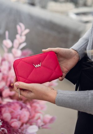 Hands holding a red quilted wallet with a silver "W" emblem, against a blurred background of pink flowers and stone pavement.