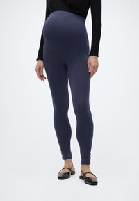 Trudnica koja nosi mornarsko plave trudničke tajice sa zlatnim gumbima kod gležnjeva i crne ravne cipele, stoji na bijeloj pozadini.