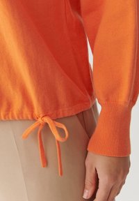 Pomarańczowy dziergany sweter z prążkowanym wykończeniem u dołu, ozdobiony marszczonym sznurkiem w pasie i dopasowanymi prążkowanymi mankietami. Widoczna miękka tekstura.