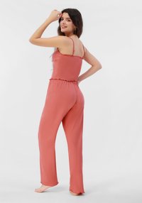 Mono de color rosa con tirantes de espagueti, tejido texturizado, cintura elástica y piernas acampanadas, con bordes rizados en el dobladillo.