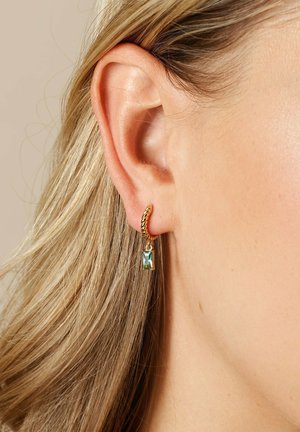 Goldene Creolen mit einem gedrehten Design, die einen rechteckigen blaug Grünstich-Gemstein-Anhänger zeigen. Nahaufnahme am Ohr für Details.