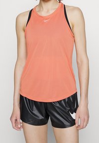 Top sportivo Nike di colore corallo brillante con una superficie liscia e scollatura rotonda, abbinato a pantaloni corti neri lucidi con un dettaglio del logo bianco.