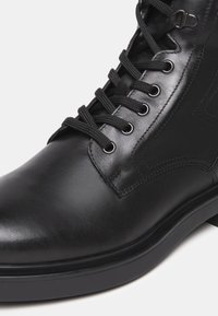 Botín de cuero negro con punta redonda, cordones negros pasados por ojales de metal y una suela baja de goma.