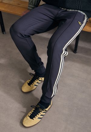 Personne portant un pantalon de survêtement noir avec des bandes blanches sur les côtés et des baskets jaunes, assise sur un banc en bois sur un sol gris.