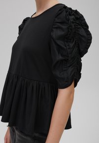 Svart topp i mjukt tyg, med en figurnära bodice och en utsvängd peplum samt voluminösa, rynkade puffärmar. Enkel rund halsringning.