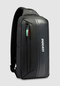 Sac à dos rectangulaire noir en matériau durable ; présente une texture lisse, une poche avant zippée et des rayures grises discrètes avec un détail de logo.